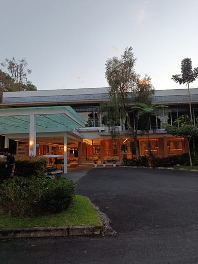 The image depicts the exterior of a modern building with a glass-covered entrance canopy. The building features large windows, with interior lighting visible. The scene appears to be set in Durin Serugun, Indonesia, as indicated by the context. In the foreground, a man in a dark uniform is standing near a lush green hedge. Adjacent to the hedge, a small, white electric cart is parked. The ground is paved with asphalt and surrounded by neatly maintained grass and shrubbery. Several trees, including one with thin, wispy branches and another with fern-like fronds, are situated in front of the building, casting shadows. The sky is overcast with hints of a sunset, suggesting it is either early morning or late evening. The overall impression is one of a tranquil, well-maintained outdoor space connected to a contemporary structure.
