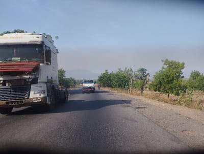 The image shows a paved road in Lakweme, Nigeria (9.706, 11.432). A large white semi-trailer truck with the words “FINMARETI” in yellow on the side is parked on the right side of the road. A smaller white truck is traveling towards the camera in the distance. The road is partially obscured by the parked truck and some trees. The vegetation is sparse and dry on both sides of the road. The sky is a clear blue and the sun is shining. The road is mostly empty. The image appears to be taken from inside a vehicle traveling along the road.