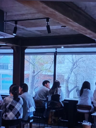 The image depicts the interior of a commercial establishment, likely a cafe or restaurant, situated in Yangiabad, Uzbekistan. The scene focuses on a counter area equipped with high stools, positioned directly in front of large windows. The ceiling is exposed concrete, featuring dark wooden beams and a track lighting system with two black directional spotlights.
Multiple individuals are seated along the counter, predominantly with their backs to the camera, looking out the windows. In the immediate foreground, a person on the left wears a grey and white argyle patterned sweater. Next to them, a person wears a light grey top with a ruffled hem, over blue jeans. Further down the counter, a male individual is visible in side profile, wearing a light grey long-sleeved shirt and gazing out the window. Adjacent to him, a female individual with long dark hair is seated, wearing a light-colored top, also facing the window. Another female individual, wearing a white long-sleeved shirt, is seated further to the right with her back to the camera.
Through the windows, an outdoor urban environment is visible, characterized by deciduous trees with some foliage and a multi-story building featuring exterior air conditioning units. The natural light from outside illuminates the scene, contrasting with the more subdued interior lighting.