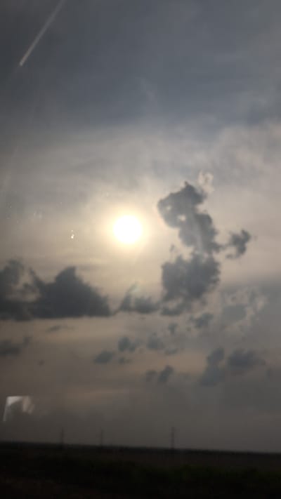 The image is a blurry, overexposed photograph of the sky. The sun is visible as a bright, blown-out circle in the upper center of the frame, surrounded by diffuse light. Dark, indistinct clouds are scattered across the sky, some appearing as dark masses and others as lighter, hazy shapes. In the upper left corner, a faint, bright streak suggests a contrail from an aircraft or a light reflection. The bottom of the image shows a dark, out-of-focus strip representing the horizon and a field, with faint vertical lines that could be distant poles or structures. The overall impression is of a cloudy, possibly hazy, sky with the sun breaking through. There are no discernible people, specific objects, or actions to describe beyond the atmospheric conditions. The location in Borcea, Romania, is not visually identifiable from the image itself.