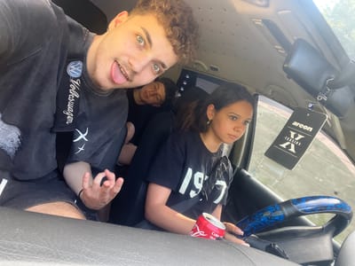 The image shows four young people in a car. The person in the foreground, on the left, is taking the selfie. They have curly brown hair and are sticking their tongue out. They are wearing a dark t-shirt with a Volkswagen logo on it. To their right, a person with their eyes closed is leaning on the seat. Further to the right, a young woman with dark hair is visible, wearing a black t-shirt with the number "1997" on it. Her left hand is resting on a red can of Coca-Cola. Another person's arm is visible on the far right. The car's interior is visible, including a steering wheel with a blue and black patterned cover and an air freshener hanging from the rearview mirror. The scene appears to be in Vlahovo, Bulgaria, as indicated by context provided with the image. The lighting suggests it is daytime.