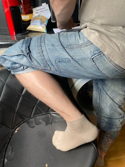 This image depicts a close-up view of a person's legs and lower torso, seated on a black chair with diamond-patterned upholstery that shows signs of wear and tear. The person is wearing light-wash denim shorts, a light-colored t-shirt, and beige ankle socks. A tattoo, primarily in shades of blue and black, is visible on the lower right leg. In the background, on a countertop, are several objects, including a red coffee machine, a yellow box with a barcode, and a small plastic bag with Estonian text and an image of a pipe. A white piece of paper with text, also in Estonian, is attached to a dark object, possibly a cabinet. The overall environment appears to be indoors, with hints of a kitchen or laundry area due to the visible coffee machine and what appears to be the top of a washing machine. The image appears to be taken in Tartu, Estonia, as indicated by the text on the paper and packaging.