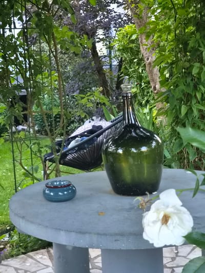 A large, dark green glass bottle with a glass stopper stands on a round, grey concrete table in a garden. To the left of the bottle is a small, blue ceramic jar with a red rim. In the foreground, to the right of the bottle, a wilting white rose is visible. Behind the table, a black woven chair is partially visible, filled with dark clothing and a patterned textile. The garden is lush with green foliage, trees, and a lawn. In the background, there are hints of buildings and a metal fence. The scene is likely a private garden setting in Bettembourg, Luxembourg.