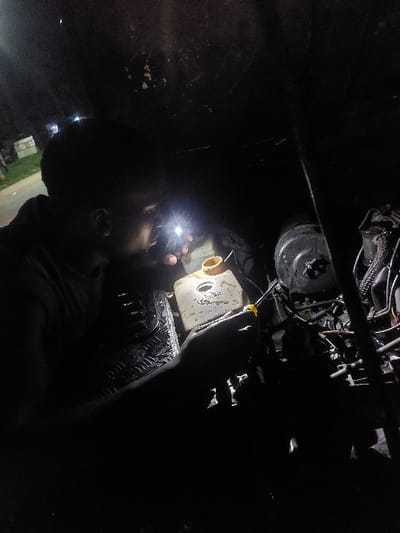 At night in Msata, Tanzania, a male individual is depicted from the left side profile, illuminating a vehicle's engine bay with a bright flashlight. His right hand holds a screwdriver with a yellow handle, positioned near a translucent, yellowish-white plastic fluid reservoir with a brown cap. The flashlight beam directly illuminates the reservoir and surrounding engine components, including various hoses and a dark, circular mechanism to the right. The individual's gaze is directed towards the illuminated area. The immediate foreground shows a dark, textured surface, possibly a floor mat or part of the vehicle's interior. In the distant, dark background to the upper left, faint artificial lights are visible, indicating an external environment.