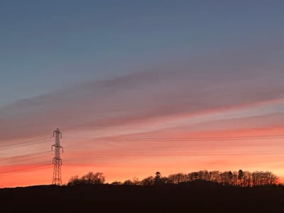 The scene, located in Muirhead, United Kingdom, displays a sunset with a sky gradient. The upper sky is deep blue, transitioning to purplish-pink with elongated cloud formations in the mid-section, and vivid orange-red hues at the horizon. A large, galvanized steel lattice-work electricity pylon is silhouetted on the left, supporting at least six visible overhead power lines that extend horizontally across the frame. The lower third of the image is defined by a dark, silhouetted landmass topped with a dense line of trees, including both bare deciduous and coniferous types. No human subjects, animals, or active events are present.