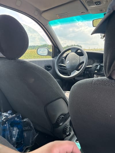 The image captures the interior of a car, possibly a Renault, viewed from the passenger side. The upholstery of the car seats is a dark grey fabric, and the headrests are visible. In the foreground, a person's hand is partially visible, with a glimpse of a fingernail. To the left, a plastic-wrapped pack of blue water bottles sits on the floor.
The dashboard features a dark grey steering wheel with a Renault logo. The car's interior is illuminated by sunlight filtering through the windshield and side windows. Outside, a rural landscape is visible, with fields of dry grass or crops and a cloudy sky. The scene suggests a journey through the countryside, possibly on a road near Opatów, Poland, given the context provided. The overall impression is one of candid travel on a sunny day.