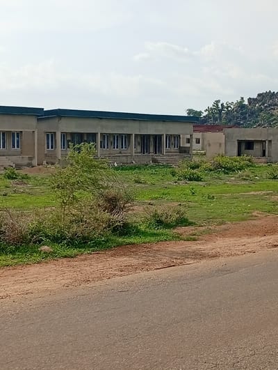 The image shows a row of unfinished concrete buildings under a cloudy sky. The buildings appear to be single-story structures with dark-colored roofs. They have numerous windows, many of which are recessed. The buildings are situated in a grassy, undeveloped area with some scattered bushes and trees. In the background, there are rocky hills with sparse vegetation. A dirt road is visible in the foreground, with an asphalt road bordering it. The scene suggests a construction site or a developing area in a rural or semi-rural environment. The city of Shuwa, Nigeria is mentioned as the location, implying this is a view of buildings and surroundings within or near that area. There are no people visible in the image.