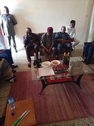 The image depicts an indoor gathering in Tambes Mile Four, Nigeria. Several men are seated on a sofa and chairs, while another man stands to the left. A glass-top coffee table is centrally placed, adorned with two bottles of wine, a bowl of snacks, several cans of what appears to be malt drink, and glasses. A small wooden table to the lower left holds a bottle of water and two knives. The room has a light-colored wall and a patterned rug on the floor.