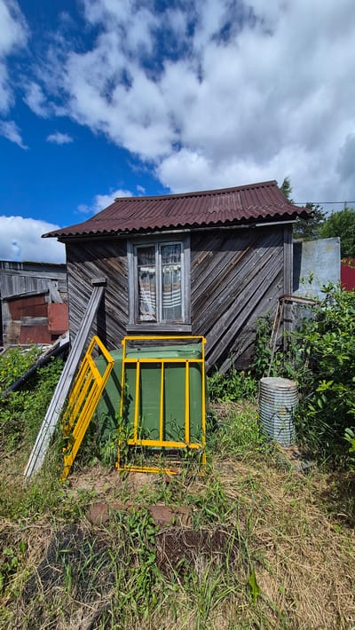 The image depicts an exterior scene in the rural settlement of Mashezero, Russia. The central focus is a weathered, dark wooden shed with a corrugated metal roof. A single window with striped curtains is visible on the shed's facade. In front of the shed, a large green trash bin is partially enclosed by a bright yellow metal frame. To the left, a section of a wooden structure, possibly another shed or fence, is leaning against the main shed. Various elements of disuse and overgrowth are present, including tall grass, weeds, a rusted metal barrel, and scattered debris. The sky above is bright blue with scattered white clouds. The overall impression is one of a neglected rural property on a clear day.
