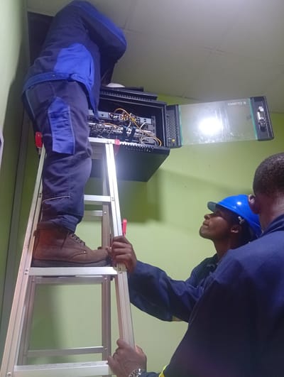 In Shiroro, Nigeria, two individuals are engaged in work related to a server rack. One person, wearing a blue and dark blue jacket with blue patches on the pants and brown boots, stands on a silver ladder. This individual is bent over and appears to be working on the equipment inside an open server rack mounted on the wall. The rack contains various cables and components, suggesting network or server maintenance.
Another person, wearing a blue hard hat and a dark blue uniform, is looking upwards towards the server rack. This individual's face is partially visible, and they seem to be observing or assisting the person on the ladder. A third individual, mostly out of focus, is also visible from the shoulders up, wearing a dark blue uniform.
The scene is indoors, with a light green wall providing a backdrop. The lighting appears to be artificial, possibly from overhead lights. A reflective surface within the server rack, possibly a glass door, shows a bright light source. Text visible on this reflective surface reads "XAMYTD S". The overall impression is one of a technical task being performed in a professional or industrial setting. The expressions on the faces are not clearly discernible, but the focused posture of the individuals suggests a task requiring concentration.