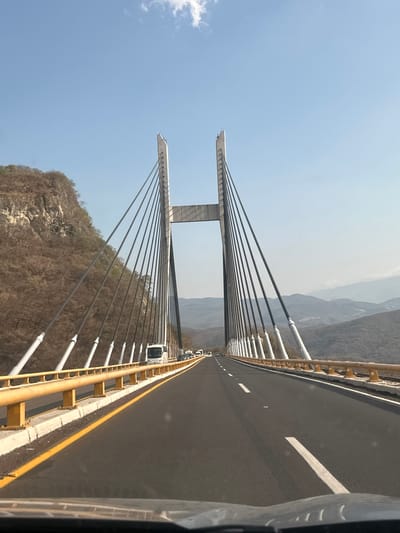 The image shows a cable-stayed bridge in Zumpango del Río, Mexico. The bridge has two large concrete towers supporting the deck with numerous black cables. A road runs along the bridge deck with a yellow and white barrier on one side. A mountain slope is visible below the bridge. Several cars are visible on the bridge. The sky is light blue with a few clouds.
