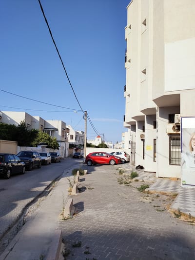 This media file captures a street scene on a clear, sunny day, likely in Tunisia, given the architectural style and the presence of an air conditioning unit with Cyrillic lettering on its control panel. The image features a row of parked cars on a street lined with multi-story buildings. The buildings are painted in light colors, with some having balconies.
On the left side of the street, several cars are parked, including a black hatchback, a black SUV, a white sedan, and another black sedan. Further down the street, a red car is parked in front of a building. The foreground shows a sidewalk made of interlocking concrete pavers. Greenery is sparse, with small bushes and weeds growing along the edges of the sidewalk and in patches of dirt.
A prominent feature of the scene is a utility pole with electrical wires running across the frame. The sky is a vibrant, cloudless blue, indicating it is daytime, likely morning or afternoon due to the shadows cast by the buildings.
On the right side of the image, a modern, pale-colored building with a textured facade stands tall. It has several small, triangular windows or recesses. An air conditioning unit is mounted on the wall of this building. A poster or advertisement is partially visible on the right edge of the frame, featuring a close-up of a person's face and some text in what appears to be French, with "TRAITEUR" and "SUP" visible. This suggests a commercial or residential area with some businesses. The overall impression is one of a quiet, everyday street in a residential or mixed-use urban environment.