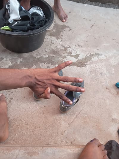 The image presents an overhead view of an outdoor scene on a concrete or paved ground, marked by several wet patches and scattered small debris like twigs.

In the central foreground, a person's dark-skinned right hand is raised, with the index, middle, ring, and pinky fingers extended upwards, while the thumb is bent towards the palm. Immediately behind the hand, a dark-colored athletic shoe or sandal with purple accents is visible.

To the left, a large, round black plastic basin is filled with various items of clothing, predominantly black and white, along with a bright green garment. On one of the black clothing items inside the basin, the text "2XL" is discernible, indicating a clothing size.

Parts of several human feet are also visible in the frame. A bare foot is partially cropped on the far left, another bare foot is seen on the far right with a purple mark near the toes, and a portion of a third foot or lower leg appears at the bottom edge. In the upper right corner, a crumpled blue object, possibly fabric or plastic, lies on the ground.

The arrangement of the basin with clothes and the wet ground suggests that laundry activities are either in progress or have recently concluded. The scene depicts a mundane, everyday activity under daylight conditions in what appears to be a practical, possibly residential, outdoor setting in Awada, Nigeria.