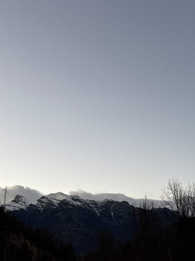 The image shows a mountainous landscape. A snow-dusted mountain range extends along the right side of the frame, its dark silhouette contrasting against a light gray sky. Bare tree branches are visible at the upper right. Utility poles are visible at the bottom right corner. The location is likely Chía, Spain.
