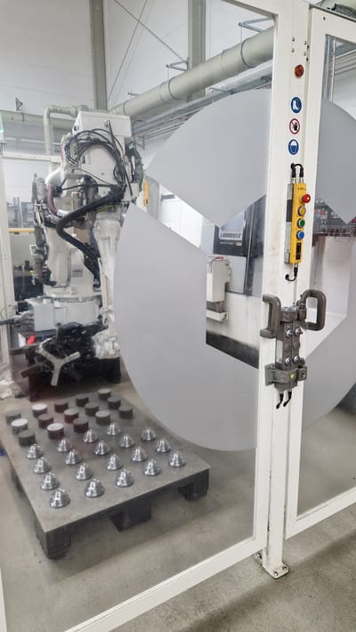 The image shows an industrial setting in Ichenhausen, Germany, likely a manufacturing or assembly plant. In the foreground, a pallet holds rows of small, metallic, cone-shaped objects, arranged neatly. To the right, a white structural element of the enclosure displays safety signage, including pictograms for eye protection, hand protection, and no entry. Below the signage, a yellow control panel with multiple buttons and a red emergency stop button is mounted. A robotic arm, white and complex, is visible in the background, connected by numerous cables and hoses. The arm appears to be positioned over a machine with a label that reads "LB3000EX II". A large, semi-transparent, curved panel, possibly part of the enclosure or a product, dominates the central and right portions of the image. The environment is industrial, with overhead pipes and a light-colored ceiling. The floor is concrete. No people are visible in the image.