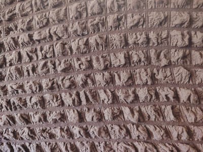 The image displays a close-up, overhead view of a textured brown fabric with a repeating pattern of embossed animal figures. The fabric appears to be made of a soft, woven material, possibly a blanket or upholstery. The animals, which resemble elephants and possibly other mammals, are depicted in profile, arranged in horizontal rows. The embossing creates a raised, three-dimensional effect on the surface of the fabric. The lighting suggests a natural, diffused light source. There is no indication of the city of Pitnak, Uzbekistan, or any environmental context beyond the fabric itself.