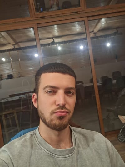 The image is a selfie of a young man with short dark hair, a beard, and a mustache. He is wearing a light gray t-shirt. He is looking directly at the camera with a neutral expression, with his lips slightly pursed. The background appears to be an interior space with large wooden-framed windows. In the reflection of the windows, several tables and chairs are visible, suggesting a restaurant or a similar establishment. The lighting in the room is dim, with visible light fixtures reflecting in the glass. There is a stroller visible in the far right of the reflection. The man is positioned in the foreground, with his face and upper body filling most of the frame. The photo appears to be taken indoors, possibly in the city of Kamenar, Bulgaria, based on the provided location information.