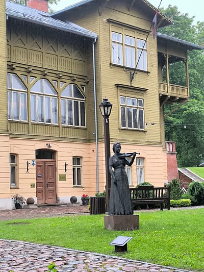 The image captures the exterior of the Kuldīga District Museum, located in Kuldīga, Latvia. The museum is housed in a two-story wooden building, painted a pale yellow on the lower level and a muted olive green on the upper level. The building features elaborate wooden ornamentation, particularly on the upper facade and the balcony.
In the foreground, a dark bronze statue of a woman playing a violin stands on a stone pedestal. She is depicted in a long dress. To the right of the statue, a wooden park bench is visible on a grassy lawn. The area in front of the museum is paved with cobblestones.
Two large spherical stone planters flank the entrance, each containing flowers. The entrance itself is a double wooden door. Above the door, a sign reads "MUZEJS," which is Latvian for "Museum." A small directional sign with an arrow and a wheelchair symbol is located to the left of the entrance.
A lamppost stands to the left of the statue, and string lights are visible strung across the scene. In the background, mature trees with green foliage are present, suggesting a park-like setting. The sky appears overcast, and there are visible streaks of rain on the building and in the air, indicating wet weather.