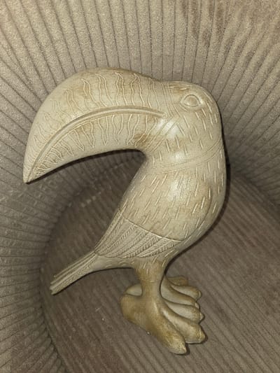 The image presents a single, static, light-toned sculpture depicting a stylized avian figure, likely a toucan or similar large-billed bird. The sculpture, which appears to be crafted from a material resembling carved stone, resin, or plaster, exhibits an overall off-white to light beige coloration with visible, irregular brownish-yellow patination or staining, particularly pronounced in recessed areas and on its base. Key carved details include a prominent, elongated, downward-curving beak adorned with linear and wavy patterns, a defined circular eye on the head, and intricate feather-like carvings across the body and wings. The bird stands on two short, stout legs culminating in splayed, paddle-like feet that form a stable base. The object is positioned on a light brown, vertically ribbed textile surface, consistent with upholstered furniture. No other objects or individuals are present. The scene is illuminated by an unseen light source, generating soft shadows that highlight the textures of both the sculpture and the background fabric. This image was captured in an unspecified interior location within Blainville-sur-Orne, France.