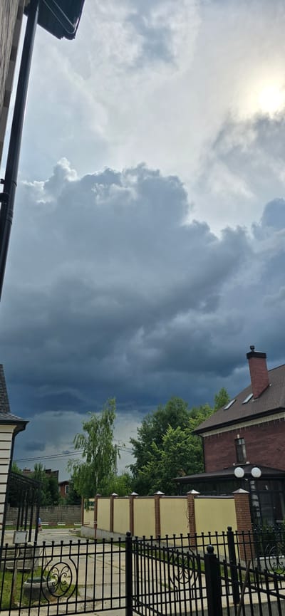 The image depicts a dramatic sky filled with dark, looming storm clouds, suggesting an impending downpour. The sun is visible in the upper left corner, partially obscured by lighter clouds, indicating it might be late afternoon or early evening. The scene is a residential area, likely in Russia, as indicated by "Русская Деревня-2 Кп".
In the foreground, the dark metal railing of a fence is prominent, with decorative scrollwork. Beyond the fence, a yellow building with brick accents and a dark-tiled roof is visible. A prominent brick chimney rises from the roof of a red-brick building adjacent to the yellow one. Green trees with lush foliage are scattered throughout the scene, some appearing windswept, further reinforcing the stormy weather. Power lines are also visible crisscrossing the sky in the distance. The overall impression is one of nature's power and the calm before a storm in a suburban or semi-rural setting. There is no visible text in the image.