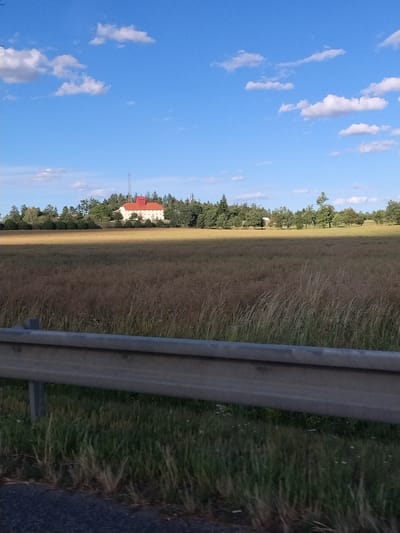 The image displays a rural landscape under a blue sky with scattered white clouds, taken from a roadside. In the foreground, a metal guardrail with support posts runs horizontally across the bottom of the frame, separating the road from a field. The field is covered in dry, tall grass and some sparse greenery. In the middle ground, a line of trees borders the field, and beyond them, a building with a red roof is visible, possibly a farmhouse or estate. A tall communication tower stands behind the building. The background consists of more trees and the sky. The scene is static and appears to have been captured during daylight. The location is identified as Drhovle Zámek, Czechia.