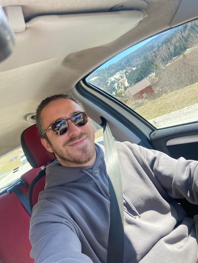 A man with a beard and sunglasses is sitting in a car, wearing a gray hoodie and a seatbelt. The car is passing through a rural landscape, possibly near Sedlačka gora, Montenegro, as seen through the window. The landscape includes bare trees and some buildings.