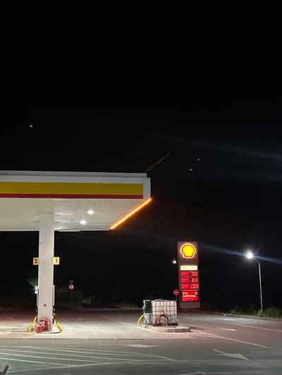 The image depicts a Shell gas station at night, located in Osenets, Bulgaria. The gas station features a canopy with yellow and red trim, illuminated by overhead lights. A white support pillar for the canopy displays the numbers "2" and "1" in yellow squares. To the left of the pillar, a red fire extinguisher and bucket are visible.
To the right of the canopy, a tall, illuminated Shell sign displays fuel prices: "1.56" for "Unleaded," "1.60" for "Diesel," and "0.68" for "V-Power." A street lamp illuminates the area. In the foreground, asphalt pavement is marked with white arrows indicating traffic direction. A white cage-like structure with hoses and a red canister is positioned near the fuel pumps. The background consists of a dark, rural landscape with sparse vegetation and trees. Several faint, blurry lights are visible in the dark sky.