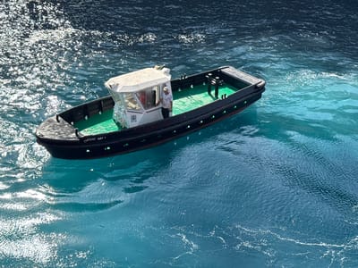 A tugboat with a white cabin and a green deck is on dark blue water. A man in a white shirt and a cap is standing in the doorway of the cabin. The boat has "CHEMAR ARZ 1" written on its side. The water is choppy and reflects the sunlight, creating bright white highlights. The water transitions from dark blue to a lighter turquoise hue in the background. The scene is observed from a high angle.