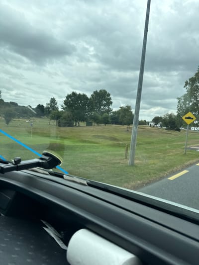The image, taken from inside a vehicle, shows a view through the windshield looking out onto a grassy landscape, likely a golf course, under a cloudy sky. The foreground features a portion of the vehicle's dashboard, including a suction cup mount. A roll of white material, possibly paper towels or toilet paper, is visible on the dashboard. Blue tape or ribbon is also seen running across the windshield.
The middle ground displays rolling green hills with scattered trees and a few structures in the distance, including what appear to be mobile homes or caravans. A tall, grey pole, likely a lamp post, stands prominently in the middle right of the frame. A yellow diamond-shaped sign with a black symbol indicating a speed bump is also visible on the right side of the road. A paved road with yellow lane markings runs along the right edge of the image. The overall environment suggests a rural or semi-rural setting, possibly near recreational grounds. There are no discernible individuals actively engaged in any actions. The location is identified as Curragh Edge, Ireland.