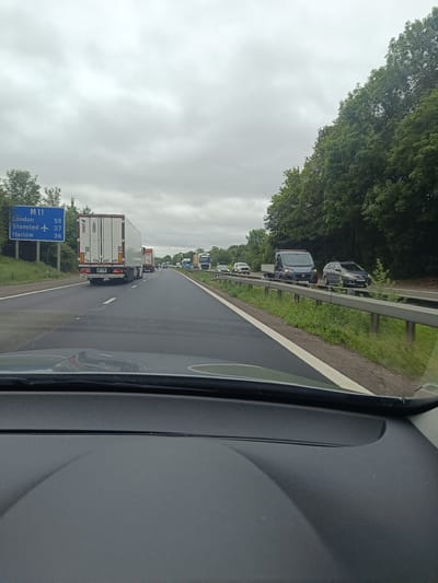 The image captures a view from a vehicle driving on a highway, likely the M11, based on the road sign. The sky is overcast, and the road is flanked by lush green trees on the right. Several vehicles, including trucks and cars, are visible on the road, suggesting moderate traffic. A blue road sign indicates directions to "M11 London," "Stansted" with an airplane icon, and "Harlow." The foreground shows the interior of the vehicle, with the dashboard visible.