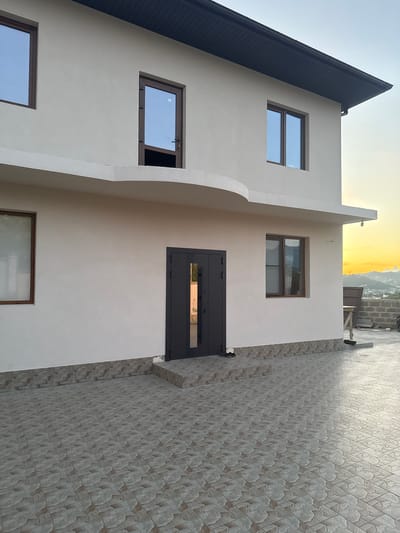 A modern, two-story house with a beige stucco exterior and a dark gray roof is situated in Verkhnevesyoloye, Russia, with a distant view of a hilly landscape at sunset. The house features several windows with brown frames, reflecting a pale blue sky. A dark gray double door is visible on the ground floor, with a small set of steps leading up to it. The ground in front of the house is paved with light gray, patterned tiles. The overall impression is one of new construction or renovation.