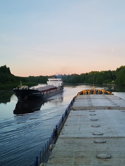 This image captures a tranquil twilight scene on a river or canal in Volokov Most, Russia. The photograph is taken from the deck of a large vessel, likely a barge, looking forward and observing another ship passing. The foreground vessel's deck features numerous circular hatches and a blue safety railing along its side.
A bulk carrier cargo ship with a dark hull and a white superstructure is seen moving in the opposite direction on the calm waterway. Its open cargo hold is filled with a light-colored material, appearing to be sand or gravel. The name "АРКАДИЙ ИВГИЕНЪ" (Arkady Ivginyev) is clearly visible in white lettering on the bow of this passing ship. It leaves a noticeable wake in the water.
Both banks of the waterway are densely forested with lush green trees, which reflect faintly in the water. The sky displays a soft gradient of colors, transitioning from light blue at the top to a gentle orange and pink hue near the horizon, indicative of either dawn or dusk. A faint, spherical object, identified as the moon, is visible in the upper right portion of the sky. The overall atmosphere is peaceful and serene, with no people visibly present.