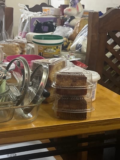 In San Antonio, Venezuela, a wooden table holds various food items and containers. In the foreground, a clear glass bowl with metal lids contains a green cloth and a metal utensil. To the right, three stacked clear plastic containers hold slices of cake. Behind these, a white plastic container with a yellow label that reads "CORRAL" is visible. A pink bowl is placed to the left of the cake containers. In the background, a large stuffed unicorn toy is visible amongst various plastic bags and boxes. A wooden chair is positioned on the right side of the frame. The lighting is dim, suggesting an indoor setting, likely a dining area or kitchen.