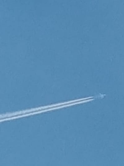 The image shows a clear blue sky with a long, white contrail from an airplane extending diagonally across the frame from the lower left to the upper right. The contrail is thin and appears to be dissipating slightly at its eastern end, suggesting the plane is moving westward. There is no discernible land or city visible in the image; it is a sky-only view.