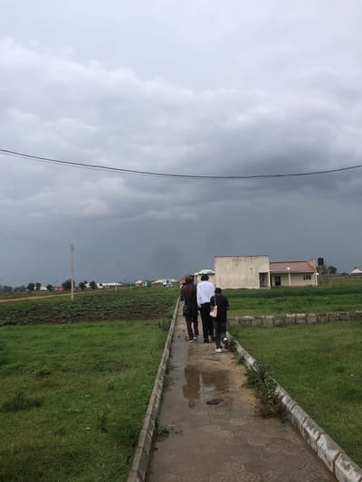 The image depicts a group of individuals walking along a wet, paved walkway. The sky is overcast with dark, stormy clouds, suggesting impending rain or recent precipitation. The scene appears to be outdoors in a semi-urban or developed area, with a neatly manicured grassy area bordering the walkway.
In the background, there are buildings, including what looks like a school or office building with a red roof and another concrete structure. Further in the distance, a few more buildings and some trees are visible, along with power lines supported by concrete poles. The ground beside the walkway is covered in lush green grass, and a section of darker, tilled earth or a field is also visible to the left.
The group of people are walking away from the camera, their figures partially obscured by the angle and the weather. One person in the foreground is wearing a white shirt, and others are in darker attire. The walkway is wet and shows reflections, indicating it has recently rained or is still raining. The overall mood is somber due to the dark sky. The location is identified as Farin Yashe, Nigeria. No visible text is present in the image.