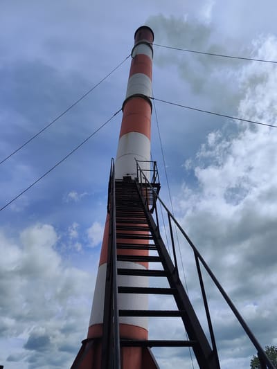 A low-angle, wide shot captures a tall, red and white striped smokestack against a cloudy sky in Milkovo, Russia. A dark metal staircase with railings ascends the side of the smokestack, disappearing out of frame. Wires are visible stretching across the sky. Smoke or steam billows from the top of the smokestack. The base of the smokestack is wider and rests on a metal support structure. The environment appears to be outdoors with some green foliage visible in the lower right corner of the frame.