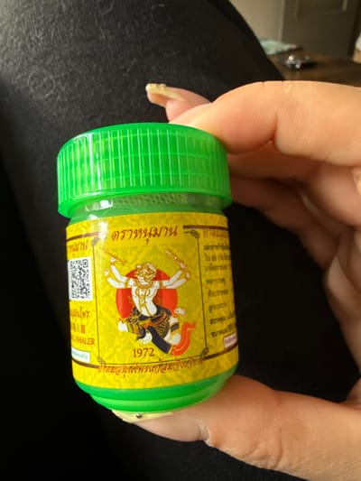 A human left hand, with light skin tone and a long, light-colored fingernail on the index finger, grasps a small, cylindrical jar. The jar features a bright green, ribbed screw-top lid and a yellow label adorned with Thai script and a red circular emblem. Within the emblem, a white figure resembling Hanuman, depicted with multiple arms wielding weapons, is visible. Below the figure, the year "1972" is printed. A QR code and the English word "INHALER" are also present on the label. The visible contents of the jar below the label are a light green substance. The background is predominantly dark, textured fabric, consistent with clothing, with a blurred lighter object partially visible in the upper right. No identifiable markers for the city of Sint-Niklaas, Belgium, are present in the scene.