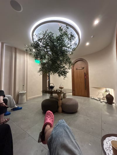 The image depicts an indoor waiting area, likely a spa or relaxation lounge. A prominent feature is a large, artificial olive tree in the center of the room, positioned under a circular ceiling light fixture. In the foreground, a person's legs and feet are visible, one foot wearing a bright pink Croc. Other individuals are seated on the floor or on ottomans to the left, their legs and feet also visible. To the right, an arched wooden door with a dark handle is set into a beige wall. Above the door, a wooden sign with Cyrillic lettering is present. A low glass table with a teapot and cups is set up near the olive tree. The floor is tiled with a light gray, speckled material. The walls are a light beige with subtle vertical paneling on the left side. The overall ambiance suggests a tranquil and modern space. The location is identified as Postol, Russia.