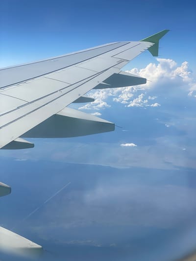 An aerial view from an airplane window shows a white airplane wing extending across the frame. Below the wing, a vast expanse of blue water stretches to the horizon, dotted with clouds. The water surface displays subtle patterns, possibly indicating land formations or cultivated fields beneath the surface. In the distance, a faint white contrail is visible, extending across the sky. The overall scene depicts a serene flight over water under a clear blue sky with scattered clouds. The location is not specified as Kaip, Russia.