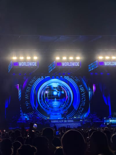 The image depicts a concert stage with a large, circular screen displaying a globe. The words "MR WORLDWIDE" are prominently displayed on illuminated signs above the screen, and also in a circular pattern around the globe. A performer is visible on stage, silhouetted against the bright lights and screen. In the foreground, a crowd of concertgoers is seen from behind, many holding up their phones to record the performance. The stage lighting is a mix of bright white spotlights and vibrant blue and purple hues emanating from the screen and decorative elements. The environment appears to be an outdoor venue or a large indoor arena, with the sky visible above the stage rigging in the upper portion of the image. The concert appears to be taking place in Lija, Malta.