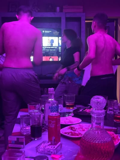 This image depicts an indoor social gathering taking place in Stolnik, Bulgaria. The scene is illuminated by pervasive purple ambient lighting.
Three individuals are present. On the left, a shirtless male with short hair stands with his back mostly to the viewer, wearing grey track pants. In the center, a female with dark, wavy hair is shown in profile, facing right, wearing a dark short-sleeved top and light-colored cargo-style pants with a blue patterned fabric section around the waist. On the right, a second shirtless male stands with his back to the viewer, wearing dark shorts or underwear. All three individuals appear to be in motion, possibly dancing or actively engaged with the environment.
In the background, a large flat-screen television is visible, displaying what appears to be a YouTube interface with multiple video thumbnails and text, some of which is in Cyrillic. The television is situated within a dark, multi-level shelving unit.
In the foreground, a rectangular table, possibly with a red and white pattern, is cluttered with various items. These include a partially consumed bottle of Absolut Vodka, a red Coca-Cola can, and a clear, decorative decanter containing a reddish liquid. Multiple drinking glasses are scattered across the table, some containing dark or amber liquids. Two white plates hold remnants of food, including white square pieces resembling cheese and other unidentified food scraps. An ashtray filled with numerous cigarette butts, an open pack of cigarettes, and discarded foil or plastic wrappers are also visible on the table surface.