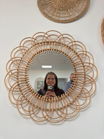 An indoor scene, located within the Eshkol Regional Council, Israel, features a prominent circular mirror with a light-toned, scalloped rattan frame mounted on a white wall. The mirror reflects a woman holding a dark red smartphone, capturing a selfie; she smiles directly into the phone, displaying long dark hair with grey streaks and dark clothing. Partially visible behind her to the right is a man with a beard, wearing a light blue collared shirt, looking left. The reflected room shows white walls, a ceiling with recessed rectangular lights, and a framed abstract artwork on a far wall. Above the mirror, a large circular woven natural fiber wall hanging is present; a second similar item is partially visible on the far right.
