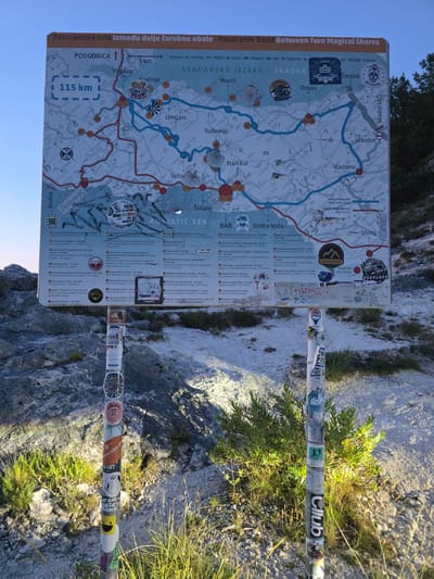 The image shows a large, detailed map mounted on a signpost in Godinje, Montenegro. The map is a panoramic route between two magical shores, featuring the Skadarsko Jezero (Lake Skadar) and the Adriatic Sea. It displays roads, cities, towns, points of interest like historical sites and monasteries, and topographical features. The signpost is situated in a rocky, outdoor environment with sparse vegetation, suggesting a mountainous or hilly area. The signpost itself is heavily decorated with numerous stickers, indicating it is a popular spot for visitors. The map is primarily in Montenegrin, with some English text. The lighting suggests it is either early morning or late afternoon.