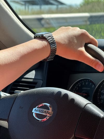 The image shows a close-up view of a person's hand wearing a silver watch, holding the steering wheel of a Nissan car. The dashboard instruments, including a tachometer and speedometer, are visible. The scene appears to be set during the daytime, with sunlight illuminating the interior of the car. The exterior is blurred but suggests a roadside environment with greenery, possibly indicating a drive through a rural or suburban area outside of Varser, Armenia.