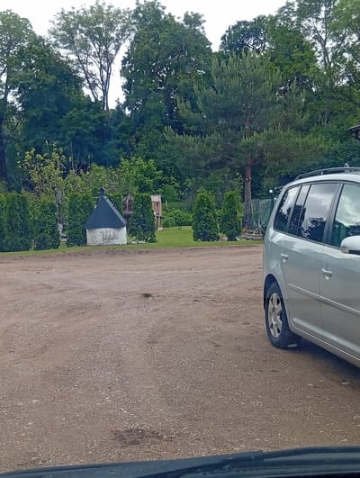 The image depicts a gravelly outdoor area in Šaukėnai, Lithuania. In the foreground, the right side of a silver minivan is visible, parked on the gravel. Beyond the minivan, a small, white, circular structure with a dark, conical roof stands in a grassy area, flanked by trimmed green bushes. Further in the background, a dense line of trees forms a backdrop, with a prominent pine tree to the right of the structure. The sky is overcast.