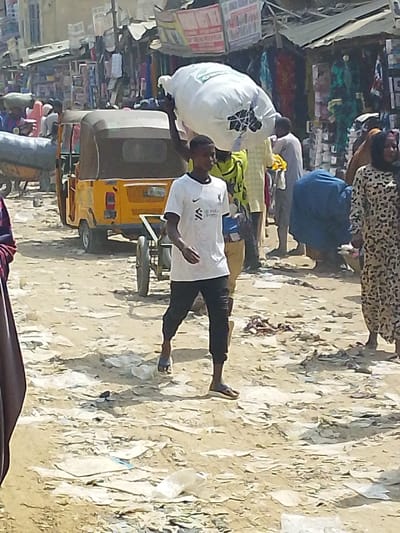 The scene is an unpaved, busy street market in Fagge D1, Nigeria, under daylight conditions. The ground is light brown dirt, heavily littered with white plastic bags and miscellaneous refuse. In the foreground, a young male, appearing to be of African descent, walks from right to left, wearing a white short-sleeved shirt with a "Liverpool FC" logo, dark trousers, and dark flip-flops. Immediately behind him, another male, also of African descent, carries a large, white, irregularly shaped sack balanced on his head, steadying it with his right hand.
In the middle ground, a yellow three-wheeled auto-rickshaw ("keke Napep") with a brown canopy is positioned towards the left, facing away from the viewer with its left side open. A simple two-wheeled handcart is visible between the walking male and the auto-rickshaw. Other individuals are present, including one person bending forward in a blue garment to the right of the walking figures, and a person in a brown and black patterned dress partially visible on the far right. The background features numerous market stalls constructed with simple frameworks and awnings, displaying various goods such as textiles and packaged items. Buildings with light-colored facades are discernible behind the market stalls.