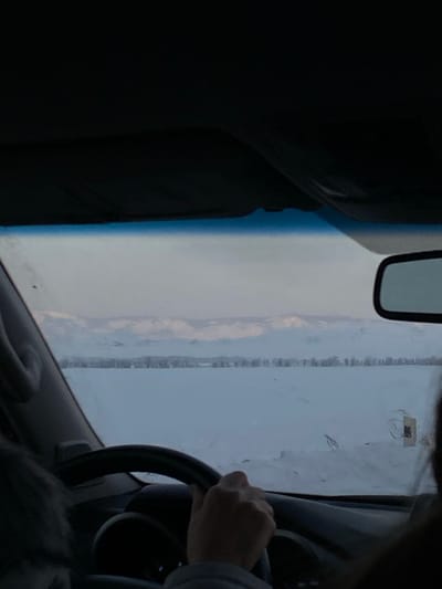 A view from inside a vehicle shows a driver with their hand on the steering wheel. The windshield is somewhat obscured, but a mountain range or hill is visible in the distance. A rearview mirror hangs from above. The car's interior is dark, contrasting with the brighter outdoor view. The scene appears to be a wintery or overcast day.