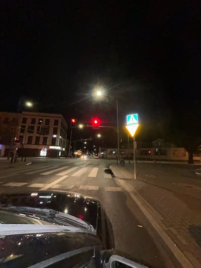 This is a nighttime photograph taken from the perspective of a vehicle at a street intersection with a crosswalk in Siedlce, Poland. The image shows a well-lit street with traffic lights showing red, indicating that vehicles should stop. A building with multiple stories and illuminated windows is visible on the left side of the street. A pedestrian crossing sign, featuring a silhouette of a person, is prominently displayed on the right. The road surface appears wet, suggesting recent rain or damp conditions. The overall scene depicts a typical urban environment at night, with streetlights and building lights illuminating the area.