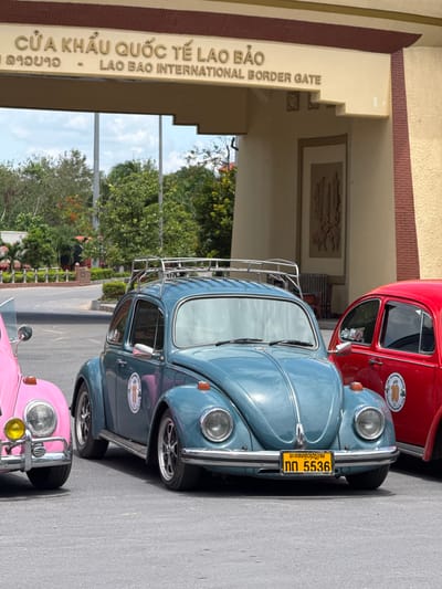 The image shows a row of classic Volkswagen Beetles parked on a paved surface. The cars are colored red, blue, and pink, and appear to be in good condition. A metal roof rack is mounted on top of the blue Beetle. In the background, there is a building with large archways and text that reads "CỬA KHẨU QUỐC TẾ LAO BẢO" and "LAO BAO INTERNATIONAL BORDER GATE." There is also text in a script that appears to be Lao, "ນະຄອນຫຼວງວຽງຈນ" and "ກຕ 5536". The scene is well-lit, suggesting it is daytime. The presence of the building and text indicates that the location is an international border gate, specifically Lao Bao. Green trees and foliage are visible beyond the building, suggesting an outdoor setting. No people are clearly visible in the image. The overall impression is of a sunny day at a border crossing, with a display of vintage cars.