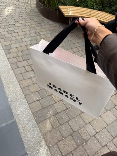 A close-up, first-person perspective image captures a person's right hand holding a white shopping bag outdoors in Las Rozas De Madrid, Spain. The hand, exhibiting manicured fingernails, is adorned with a delicate gold-toned link bracelet. The forearm is partially visible, covered by a brown jacket sleeve. The rectangular white paper shopping bag displays "ISABEL MARANT" in prominent black capital letters on its side and features black fabric handles, which the hand grips. The ground beneath is paved with rectangular, light grey interlocking pavers. A smooth, darker grey border runs along the left edge of the paved area. In the background, a curved wooden slatted bench with visible metal supports is present, positioned behind which is green foliage contained within a dark, raised planter. The scene is illuminated by natural daylight.